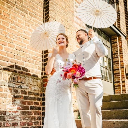 Brautpaar steht vor einer Backsteinwand und hält weiße Sonnenschirme, während die Braut einen bunten Blumenstrauß trägt und beide glücklich lächeln