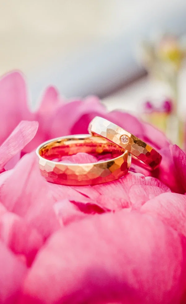 Zwei goldene Eheringe in Hammerschlag-Optik liegen auf einer pinken Blüte und strahlen im weichen Licht