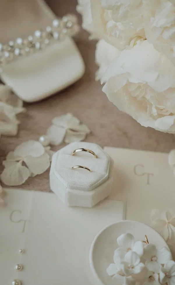 Zwei goldene Eheringe liegen in einer weißen Ringschatulle, umgeben von zarten Blüten, Papeterie und eleganten Brautaccessoires