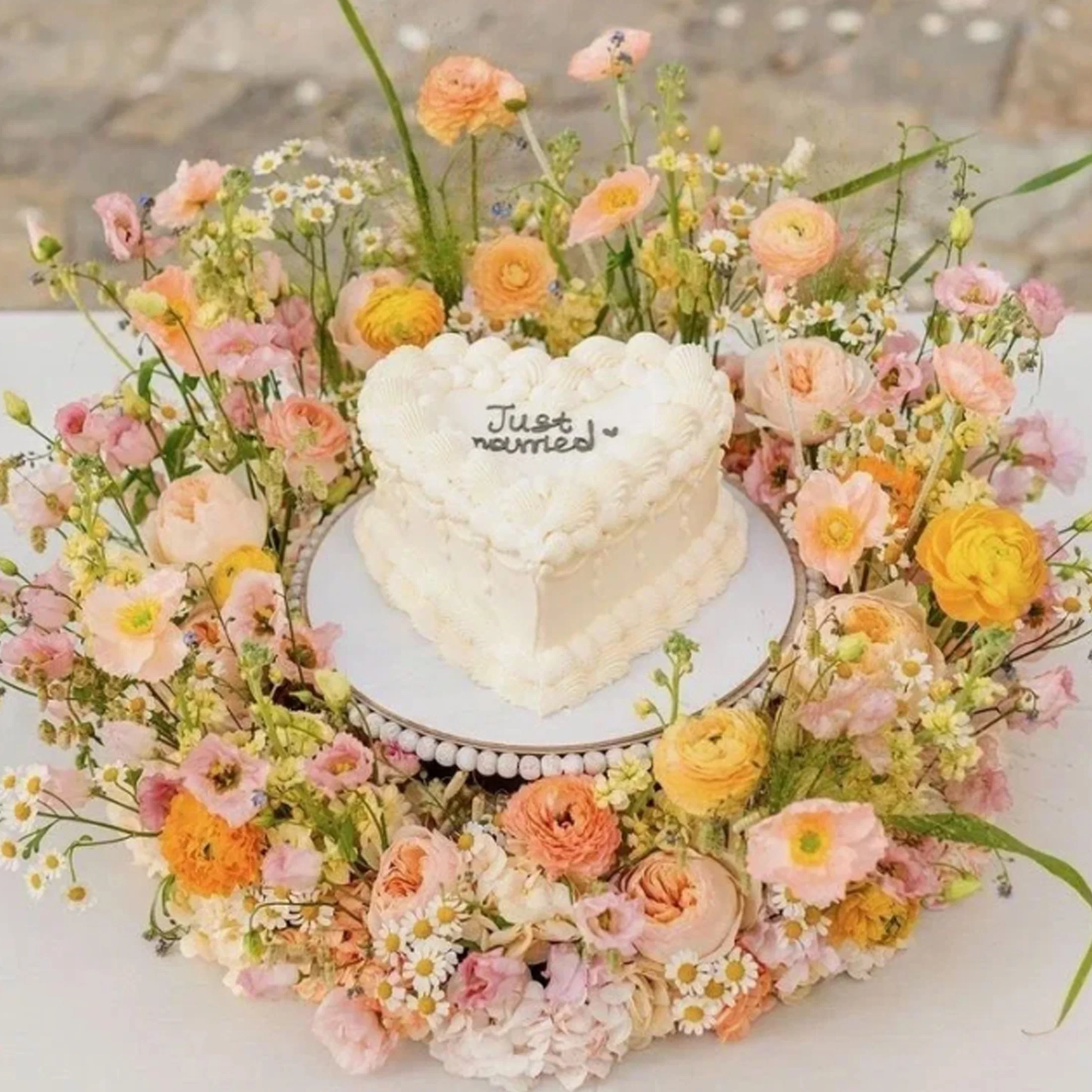 Herzförmige Hochzeitstorte mit weißem Buttercream-Frosting und „Just married“-Schrift, umgeben von zarten Blumen in Pastelltönen wie Apricot, Rosa und Gelb.