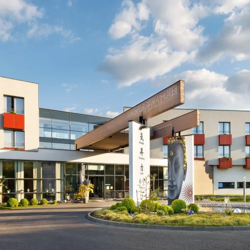 Eingangsbereich des Linsberg Asia Hotels mit moderner Architektur, Glasfassade und gepflegter Gartenanlage unter blauem Himmel