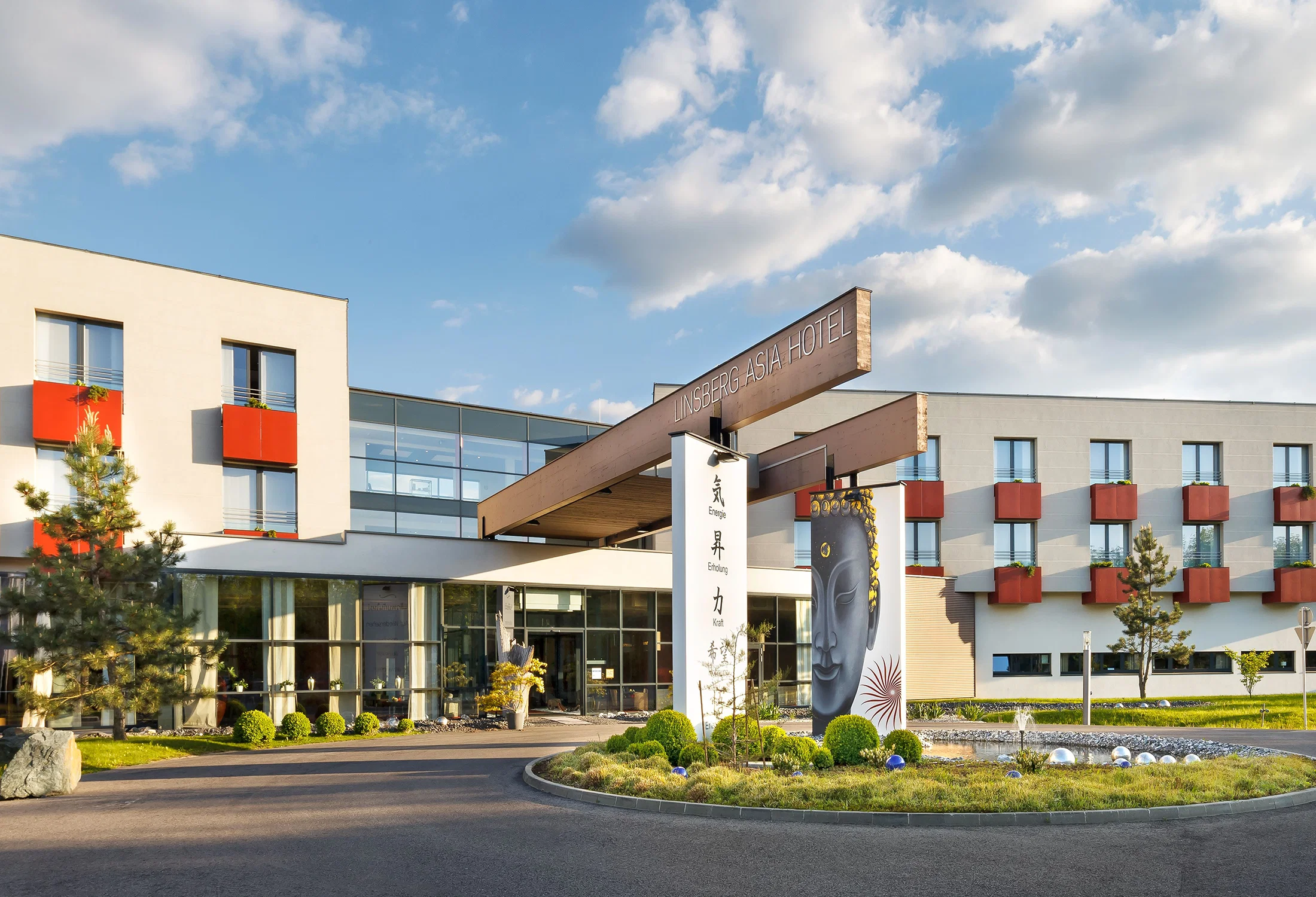 Eingangsbereich des Linsberg Asia Hotels mit moderner Architektur, Glasfassade und gepflegter Gartenanlage unter blauem Himmel