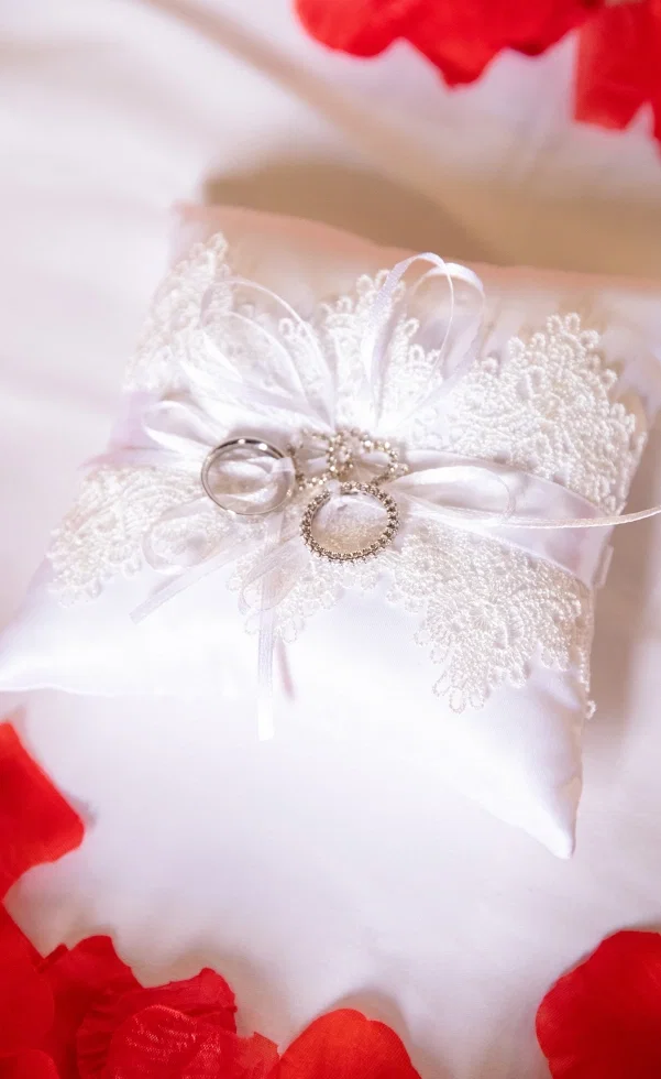 Eheringe liegen auf einem eleganten Ringkissen mit Spitze und Schleife, umgeben von roten Blütenblättern