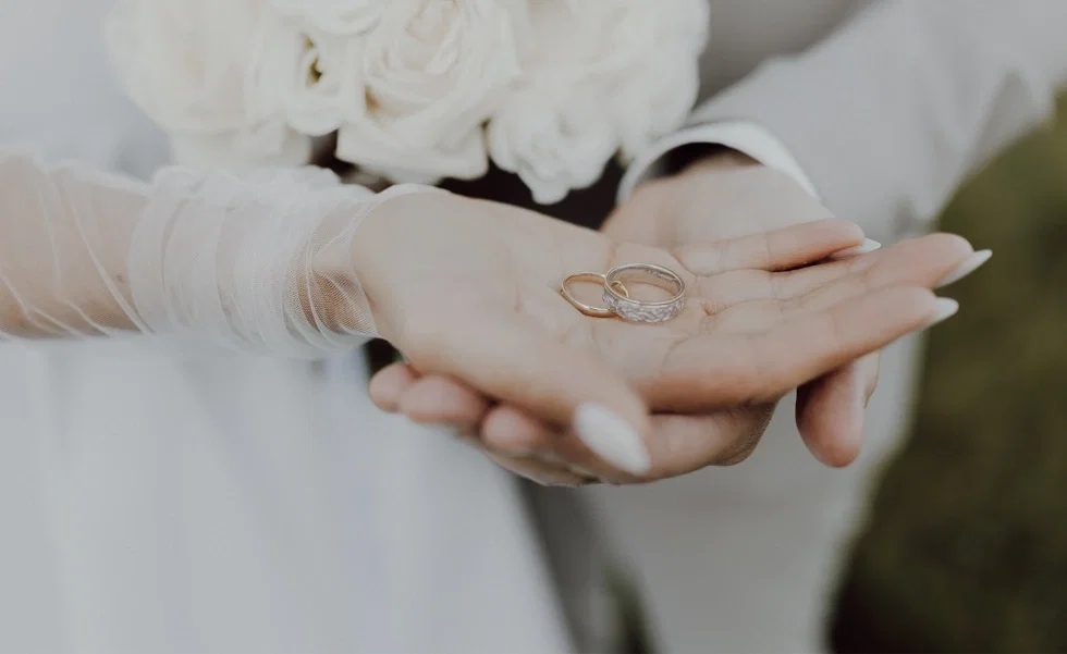 Braut hält zwei Eheringe in ihren geöffneten Händen, während im Hintergrund ihr Brautstrauß zu sehen ist