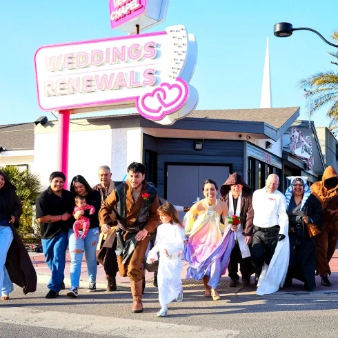 Eine fröhliche Hochzeitsgesellschaft läuft gemeinsam über die Straße vor einer Kapelle mit pinkem „Weddings & Renewals“-Schild, einige Gäste tragen fantasievolle Kostüme, während das Brautpaar im Mittelpunkt steht.