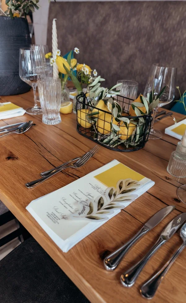Stilvolle Hochzeitstafel mit Zitronen-Deko: Holztisch mit Gläsern, Besteck, Menükarten und einem Arrangement aus Zitronen und Olivenzweigen