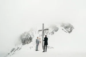 Hochzeitspaar steht bei einer freien Trauung im Schnee vor imposantem Gipfelkreuz und beeindruckender Bergkulisse.