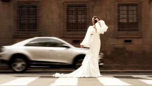 Moderne Braut im fließenden Hochzeitskleid überquert eine Straße, während ein Auto verschwommen vorbeifährt.