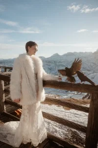Braut im Spitzenkleid und weißem Faux-Fur-Mantel steht auf einer Terrasse in den verschneiten Bergen neben zwei schwarzen Vögeln.