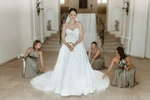 Braut steht mit ausgebreitetem Kleid im eleganten Foyer, während ihre Bridesmaids die Schleppe arrangieren.