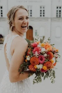 Lachende Braut mit buntem Blumenstrauß in warmen Orange- und Rosatönen vor hellem Gebäude.