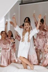 Braut und Bridesmaids feiern ausgelassen in Satin-Roben mit Champagnergläsern auf dem Bett.