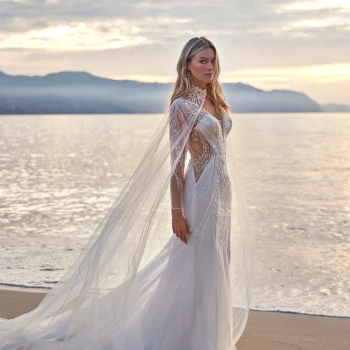 Brautkleid aus der Kollektion Deluxe by Amelie: Figurbetontes Meerjungfrauen-Kleid mit aufwendig perlensticktem, transparentem Oberteil in geschwungenen Musterlinien, kombiniert mit einem langen, fließenden Tüll-Cape mit besticktem Stehkragen. Das Cape reicht bodenlang und verleiht dem Gesamtlook eine dramatische, königliche Silhouette. Fotografiert bei Sonnenuntergang am Meer