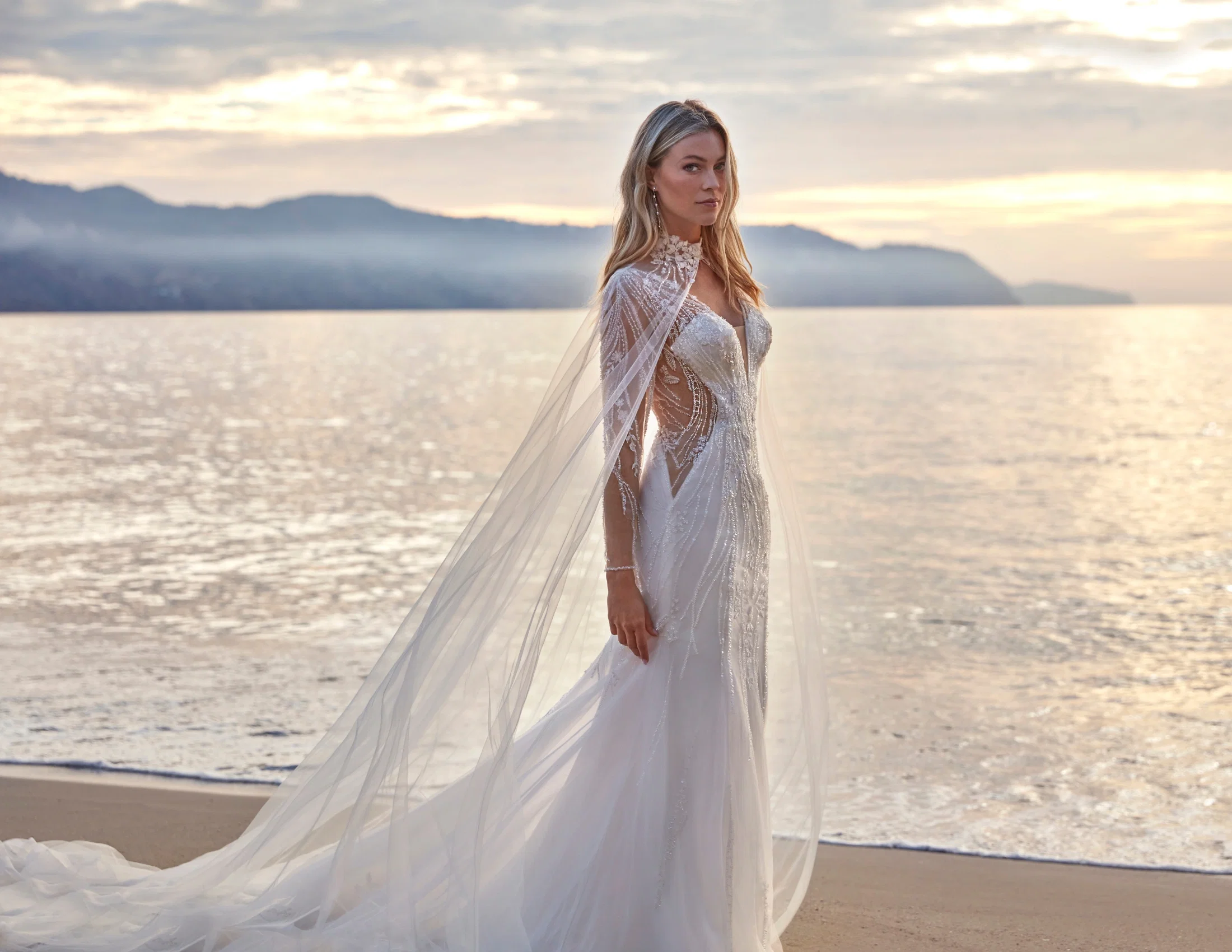 Brautkleid aus der Kollektion Deluxe by Amelie: Figurbetontes Meerjungfrauen-Kleid mit aufwendig perlensticktem, transparentem Oberteil in geschwungenen Musterlinien, kombiniert mit einem langen, fließenden Tüll-Cape mit besticktem Stehkragen. Das Cape reicht bodenlang und verleiht dem Gesamtlook eine dramatische, königliche Silhouette. Fotografiert bei Sonnenuntergang am Meer