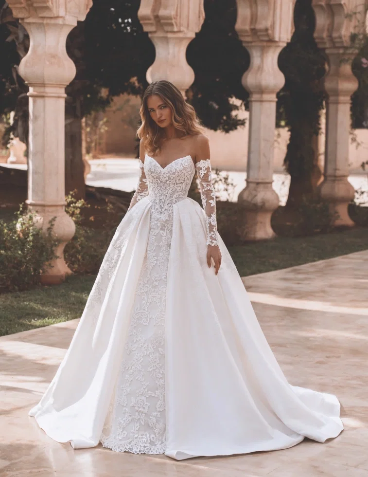 Brautkleid von Enzoani Pro Velvet: Elegantes Kleid mit besticktem Spitzen-Meerjungfrauen-Innenteil und abnehmbarem, weitem Satin-Überrock im Prinzessinnen-Stil. Off-Shoulder-Schnitt mit langen Illusions-Spitzenärmeln. Fotografiert vor einer mediterranen Säulenkulisse
