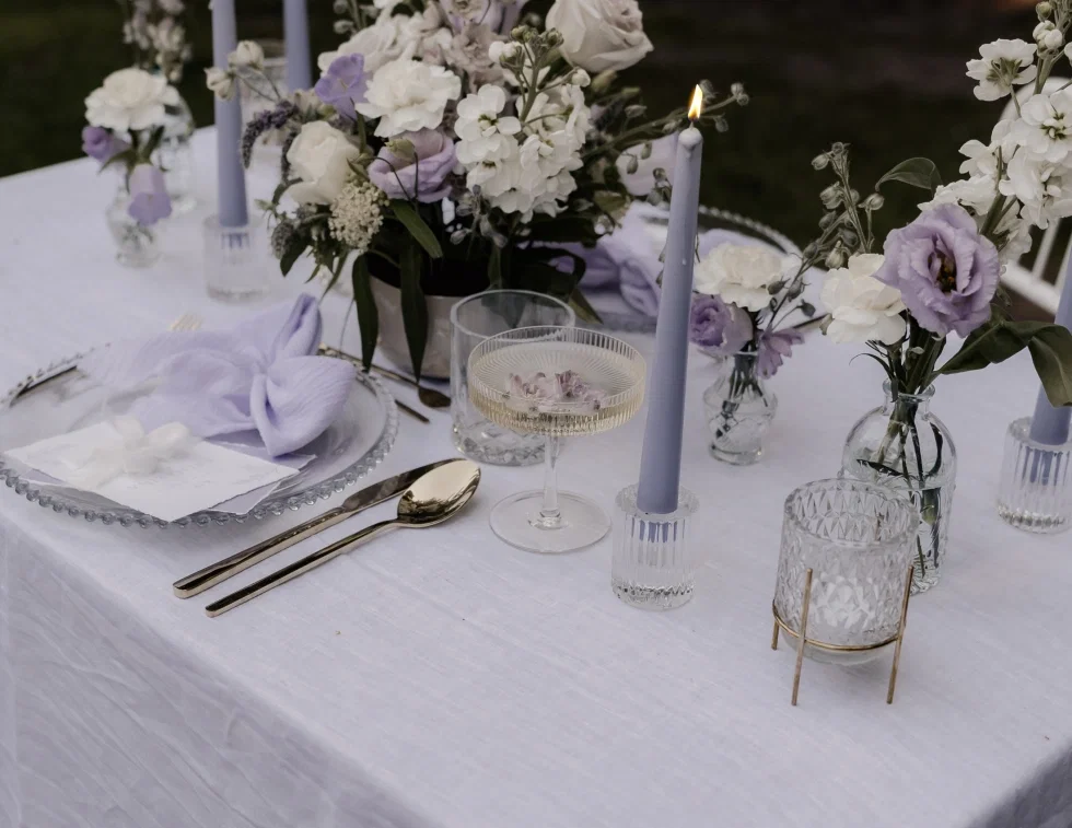 Elegant gedeckter Hochzeitstisch im Freien mit weißer Tischdecke, goldenem Besteck, gläsernem Teller mit lila Musselin-Serviette und Platzkarte, brennender lila Stabkerze, einem Champagnerglas mit Blüten sowie verschiedenen Glasvasen mit weißen und lila Blumen