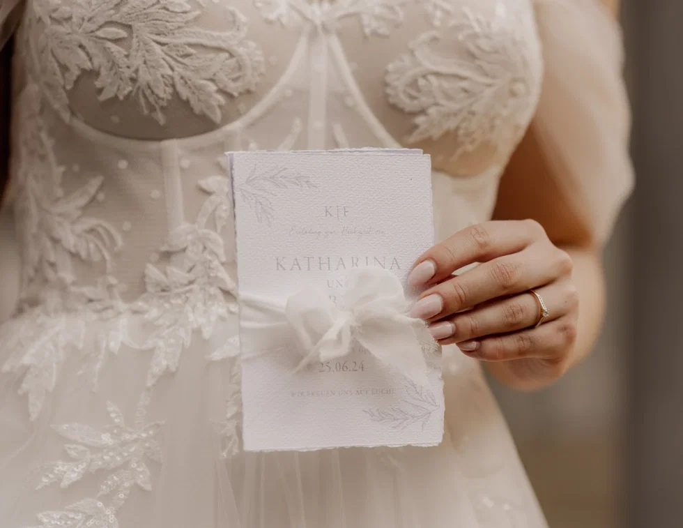 Braut in einem bestickten weißen Hochzeitskleid hält eine elegante Hochzeitseinladung mit Schleifenband in den Händen, darauf sind die Namen "Katharina" und das Datum "25.06.24" zu lesen
