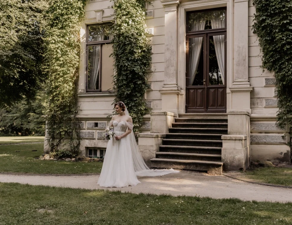 Braut in einem weißen Tüll-Hochzeitskleid mit Schleppe und Schleier steht vor einem historischen Herrenhaus mit Efeu-bewachsener Fassade und hält einen Brautstrauß in den Händen