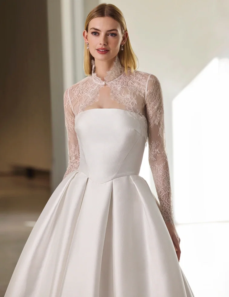 Brautkleid von Pronovias: Elegantes A-Linien-Kleid aus glänzendem Satin mit klaren, modernen Schnittlinien am Oberteil, kombiniert mit einem transparenten Spitzen-Bolero mit hohem Kragen und langen Illusions-Ärmeln aus feiner floraler Spitze. Kleiner Schlüsselloch-Ausschnitt am Halsbereich. Aufgenommen in einem hellen, minimalistischen Innenraum
