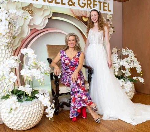 Frau im bunten Blumenkleid sitzt lächelnd auf einem Sessel neben einem Model im weißen Brautkleid, dahinter Hänsel & Gretel Schriftzug und Dekoration mit weißen Orchideen