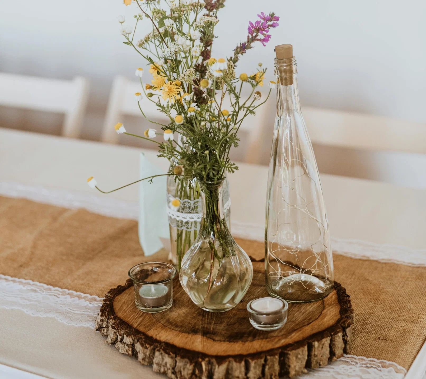 Rustikale Tischdeko mit Wildblumenstrauß in Glasvasen, Windlichtern und Deko-Flasche auf einer Baumscheibe